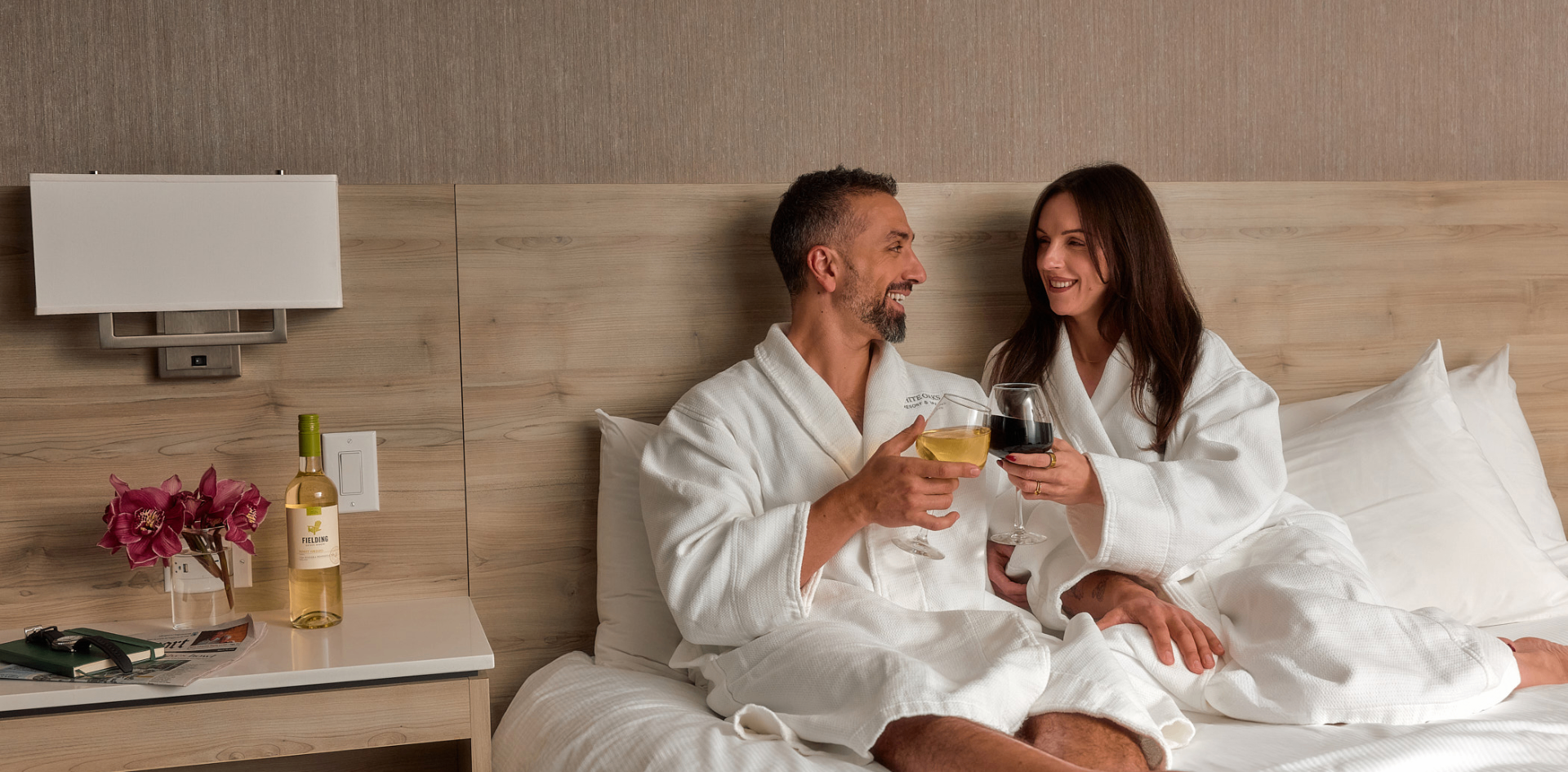 Couple in white robes toasting wine in a White Oaks Resort and Spa guest room